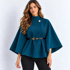 A woman modeling a teal blue cape-style top with a single decorative button at the collar, a matching black belt with a gold buckle, and black pants, set against a plain light gray studio background.