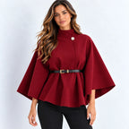 A woman modeling a deep burgundy cape-style top with a single decorative button at the collar, a black belt with a gold buckle at the waist, and black pants, set against a plain light gray studio background.