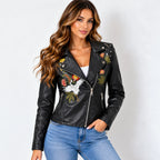 A woman wearing a black faux leather moto jacket with colorful floral and bird embroidery on the front, paired with blue jeans, against a plain light gray background.