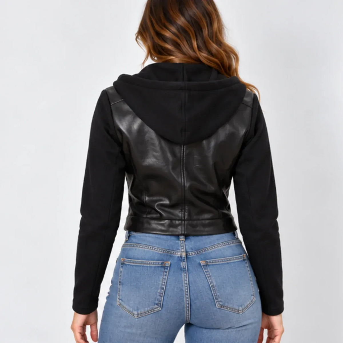 A woman viewed from the back wearing a black cropped hoodie jacket with smooth faux-leather shoulder panels and long sleeves, paired with blue jeans, against a light gray studio background.