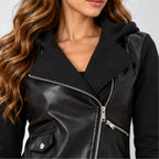 A close-up of a woman wearing a black faux-leather moto jacket with a hood and silver zipper, paired with a black hoodie underneath.