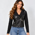 A woman modeling a black faux-leather moto jacket with a hood and asymmetrical zipper, paired with blue jeans, against a light gray studio background.