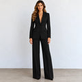 A woman modeling a black long-sleeve blazer with matching wide-leg trousers, standing against a light gray wall and concrete floor.
