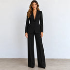 A woman modeling a black long-sleeve blazer with matching wide-leg trousers, standing against a light gray wall and concrete floor.