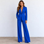 A woman modeling a bright royal blue two-piece suit with wide-leg pants and a single-breasted blazer with a deep V-neckline, standing against a light gray wall.