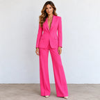 A woman modeling a bright hot pink two-piece suit with a double-breasted blazer and wide-leg trousers, standing against a light gray wall.