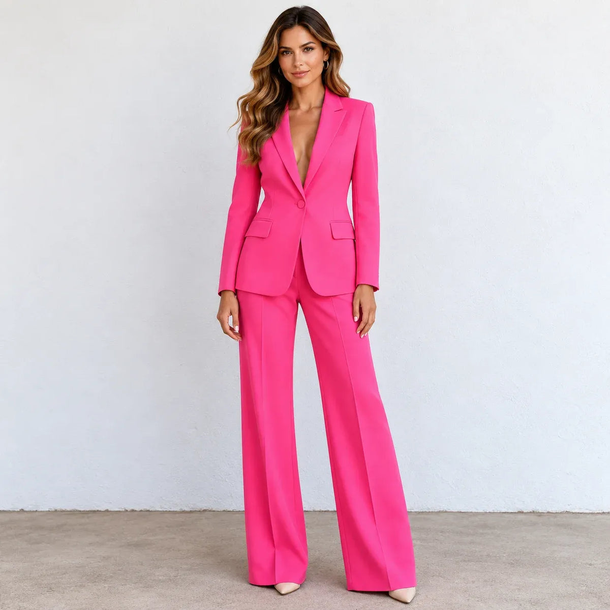 A woman modeling a bright hot pink two-piece suit with a double-breasted blazer and wide-leg trousers, standing against a light gray wall.