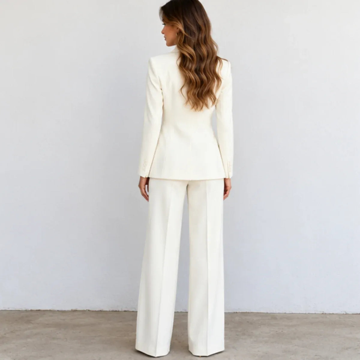 A woman viewed from the back wearing a tailored cream or off-white blazer and matching wide-leg trousers, standing against a plain light wall on a concrete floor.