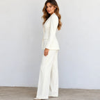 A woman modeling a white two-piece suit with a structured blazer and wide-leg trousers, standing against a light gray wall.