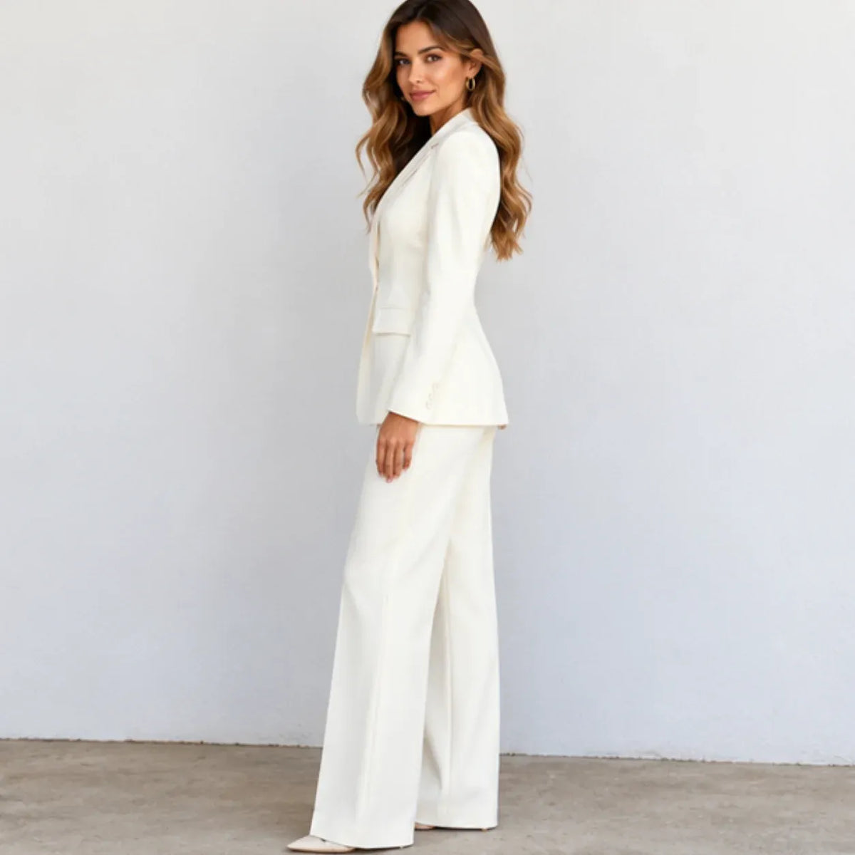 A woman modeling a white two-piece suit with a structured blazer and wide-leg trousers, standing against a light gray wall.