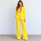 A woman modeling a bright yellow tailored pantsuit with a deep V-neck blazer and wide-leg trousers, standing against a light gray wall and concrete floor.