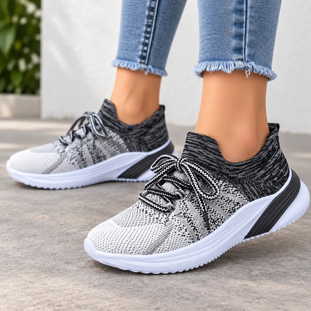 A person wearing light blue frayed-hem jeans and knit slip-on sneakers in black, white, and gray, photographed outdoors on a light concrete surface.