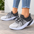 A person wearing light blue frayed-hem jeans and knit slip-on sneakers in black, white, and gray, photographed outdoors on a light concrete surface.