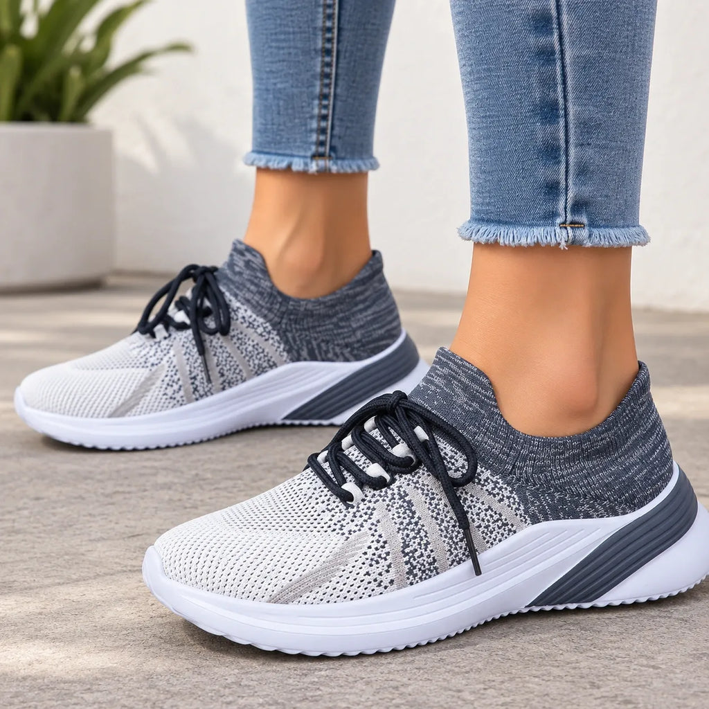 A pair of women's knit slip-on sneakers in gray and white with black laces, shown being worn with frayed blue jeans on a neutral outdoor patio background.