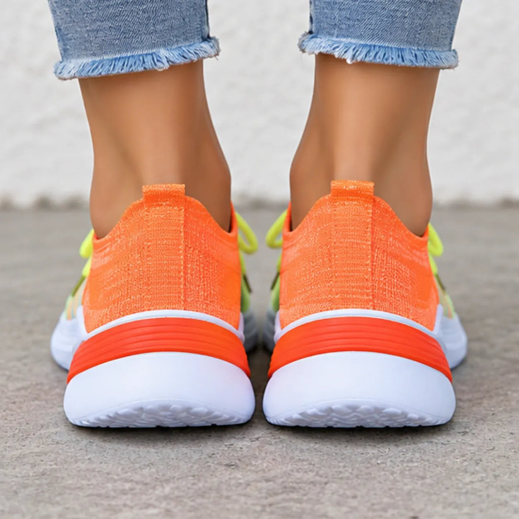 A close-up rear view of a person wearing bright orange and white athletic sneakers with light blue frayed-hem jeans, standing on a concrete surface.