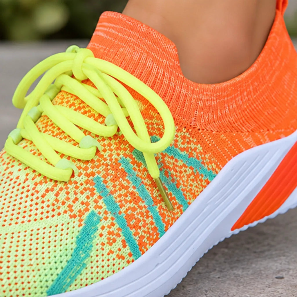 A close-up of a colorful knit athletic shoe being worn, with a bright orange and neon yellow-green upper, neon yellow-green laces, and a white sole.