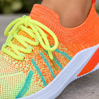 A close-up of a colorful knit athletic shoe being worn, with a bright orange and neon yellow-green upper, neon yellow-green laces, and a white sole.