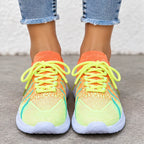 A pair of vibrant neon yellow and orange knit sneakers with lime green laces, shown on a person's feet wearing blue jeans against a concrete wall.