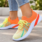 A close-up of a person wearing bright knit sneakers in orange, neon yellow, and teal, with light blue frayed-hem jeans visible against an outdoor blurred background.