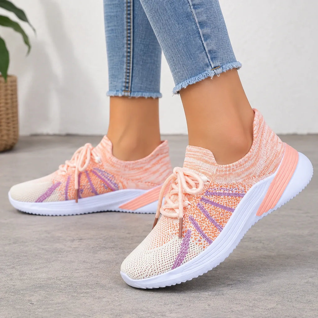 A person wearing light blue frayed-hem jeans and a pair of knit slip-on sneakers in peach, orange, and purple with white soles, standing on a gray concrete floor.