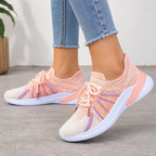 A person wearing light blue frayed-hem jeans and a pair of knit slip-on sneakers in peach, orange, and purple with white soles, standing on a gray concrete floor.