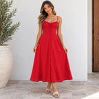 A woman modeling a red midi dress with thin straps, a sweetheart neckline, a front tie detail, and a button-up front, standing against a white wall with a potted plant on the left.