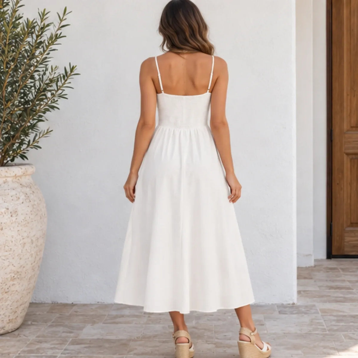 A woman viewed from behind wearing a white spaghetti-strap midi dress with a gathered waist and flared skirt, standing in front of a white wall on a tiled floor.