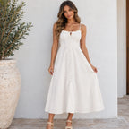 A woman modeling a white midi-length spaghetti-strap dress with a button-up front, standing against a white stucco wall next to a large potted plant.