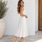A woman wearing a white spaghetti-strap midi dress with a fitted bodice and flowing skirt, paired with beige platform sandals, standing in front of a white wall.