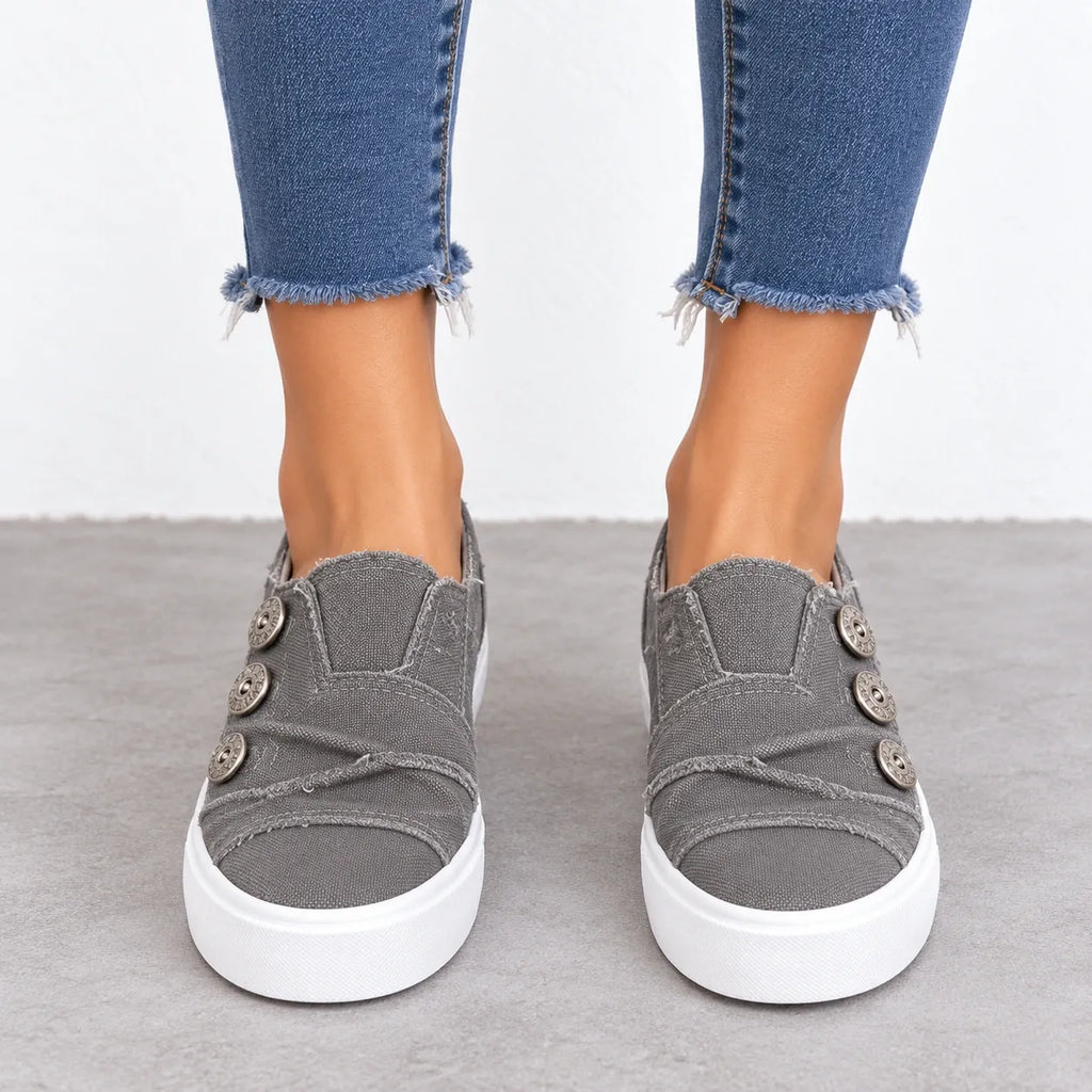 A person wearing gray slip-on platform sneakers with decorative buttons and frayed blue jeans, standing on a gray floor against a white wall.