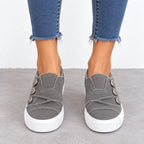 A person wearing gray slip-on platform sneakers with decorative buttons and frayed blue jeans, standing on a gray floor against a white wall.
