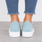 A pair of light blue canvas slip-on sneakers with frayed edges and thick white platform soles, shown on a person's feet wearing blue frayed-hem jeans.