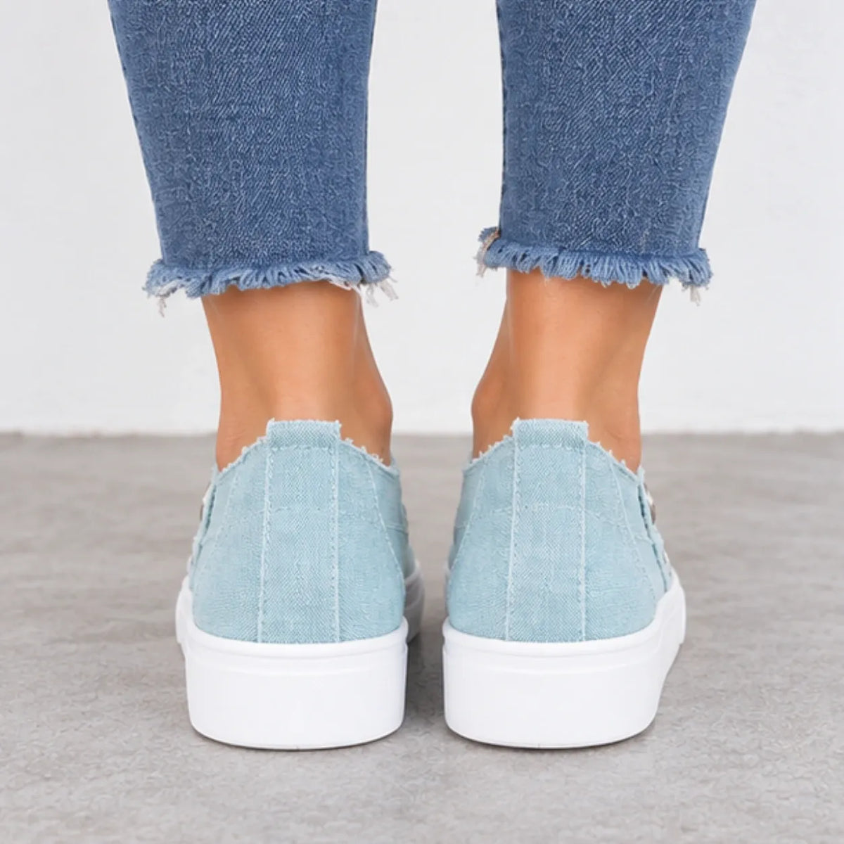 A pair of light blue canvas slip-on sneakers with frayed edges and thick white platform soles, shown on a person's feet wearing blue frayed-hem jeans.