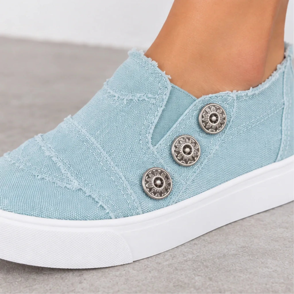 A close-up of a light blue canvas slip-on sneaker worn on a foot, featuring three decorative metallic buttons on the side and a white rubber sole, set against a neutral gray floor.