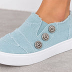 A close-up of a light blue canvas slip-on sneaker worn on a foot, featuring three decorative metallic buttons on the side and a white rubber sole, set against a neutral gray floor.