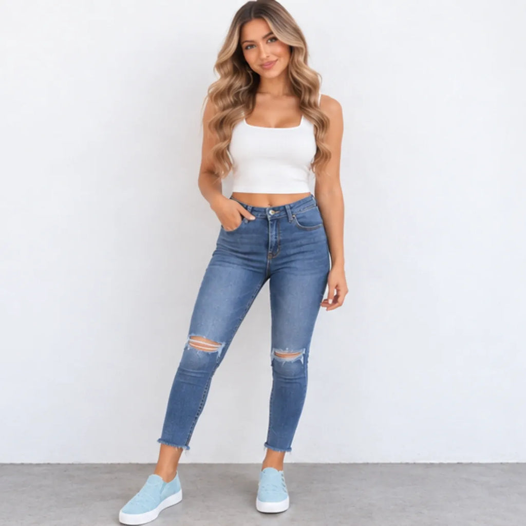 A woman modeling a pair of high-waisted, blue skinny jeans with distressed knee details, paired with a white crop top and light blue sneakers, standing against a plain white wall.