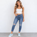 A woman modeling a pair of high-waisted, blue skinny jeans with distressed knee details, paired with a white crop top and light blue sneakers, standing against a plain white wall.