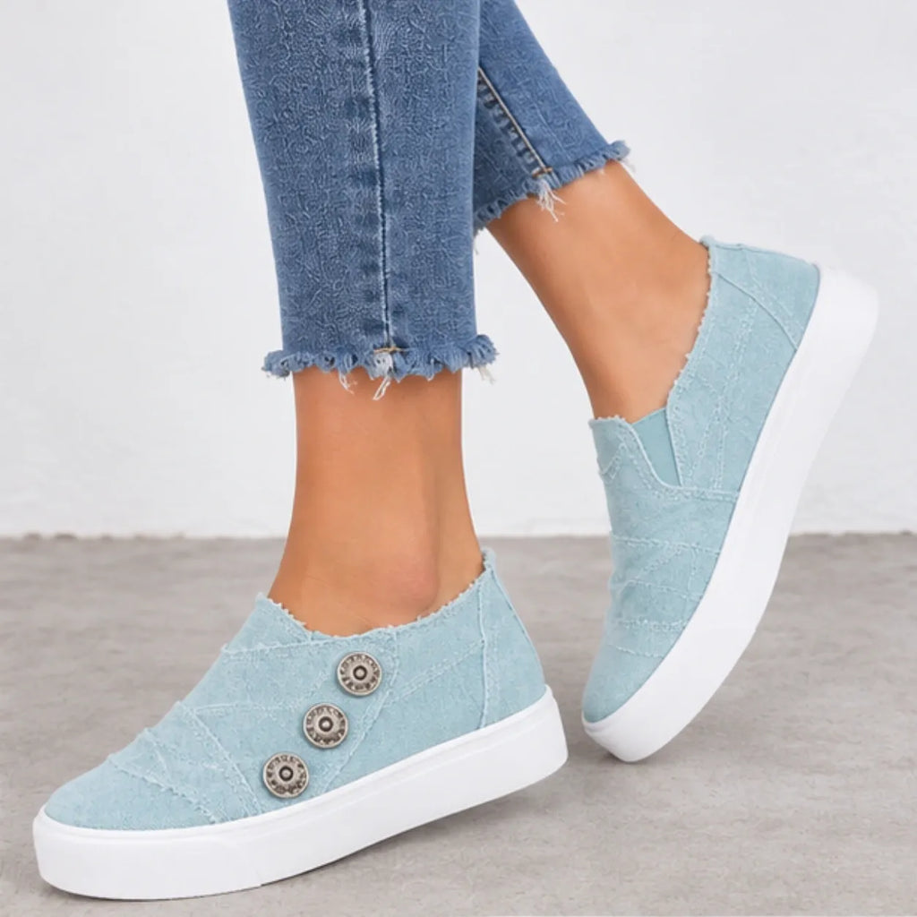 Light blue canvas slip-on sneakers with three decorative metal buttons on the side, worn with frayed blue jeans on a neutral gray floor.