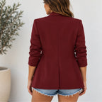 A woman shown from the back wearing a structured blazer in deep burgundy with rolled sleeves and denim shorts, standing against a light wall with a potted plant on the left.