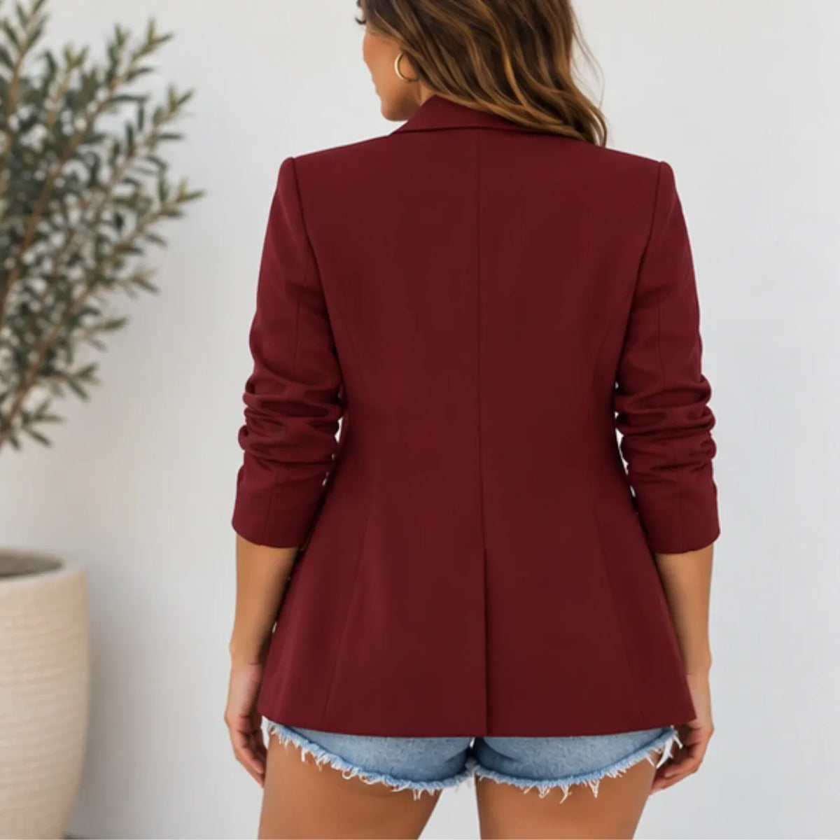 A woman shown from the back wearing a structured blazer in deep burgundy with rolled sleeves and denim shorts, standing against a light wall with a potted plant on the left.