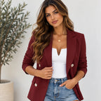 A woman wearing a burgundy double-breasted blazer over a white top and blue denim shorts, standing indoors against a light wall with a potted plant on the left.