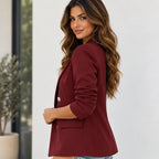 A woman wearing a tailored blazer in deep burgundy, photographed from the side and back against a clean white wall with a potted plant in the background.