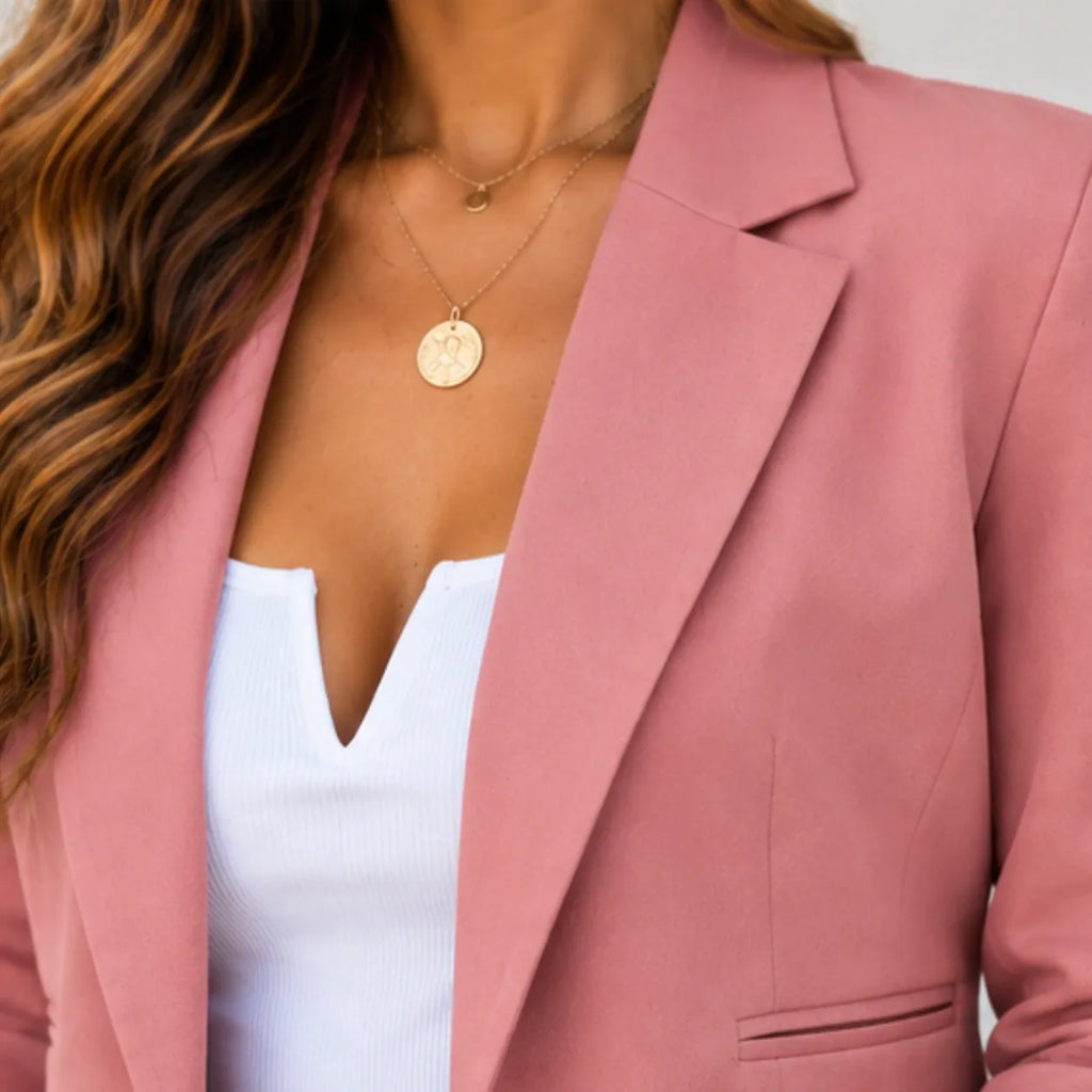 A close-up of a woman wearing a dusty pink blazer over a white ribbed top, accessorized with layered gold necklaces.