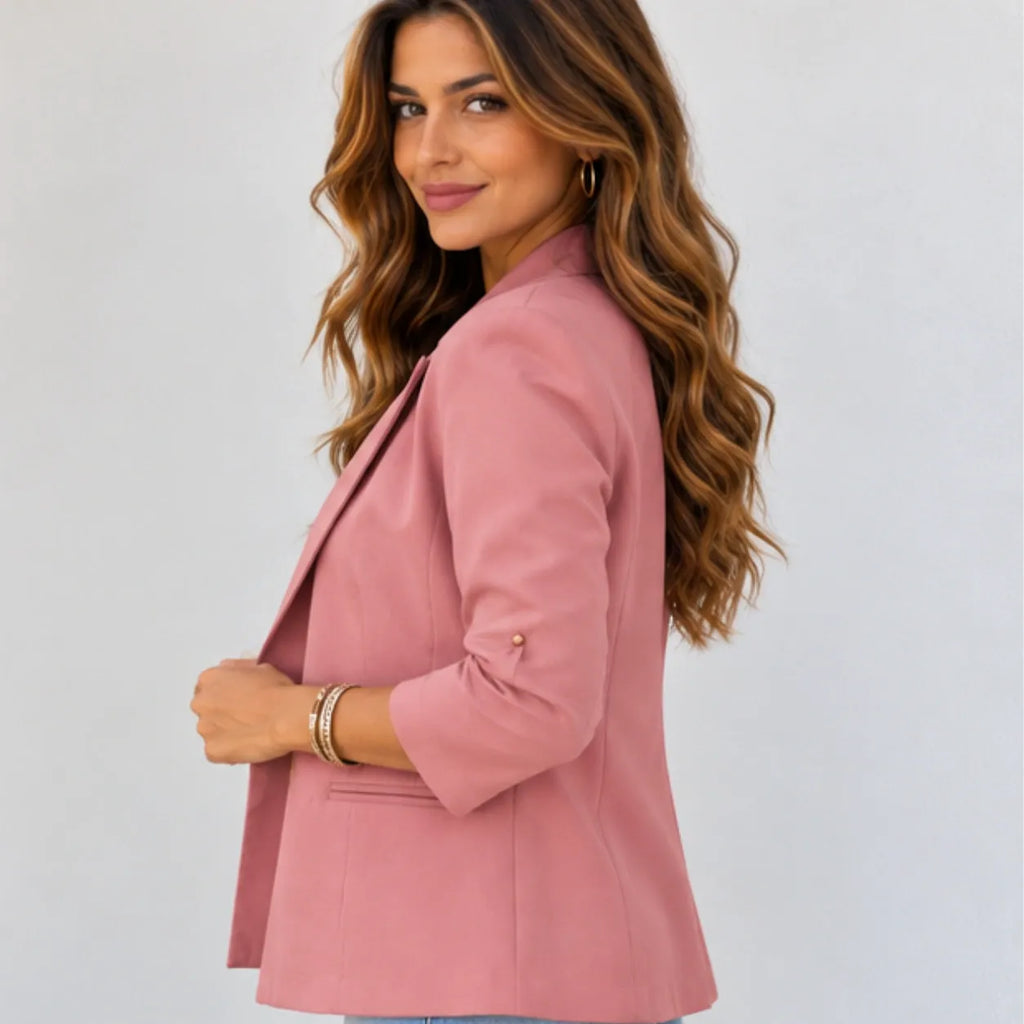 A woman with long wavy brown hair is shown from the side/back wearing a dusty pink blazer. The blazer has three-quarter sleeves, a structured silhouette, and a single visible gold button on the sleeve cuff.