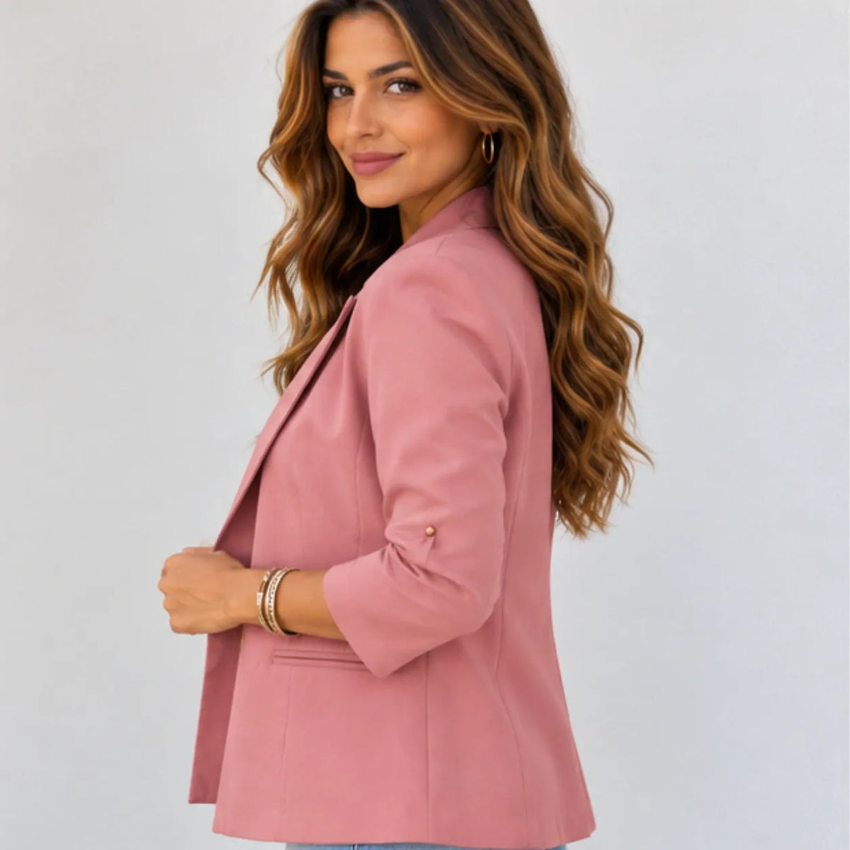 A woman with long wavy brown hair is shown from the side/back wearing a dusty pink blazer. The blazer has three-quarter sleeves, a structured silhouette, and a single visible gold button on the sleeve cuff.