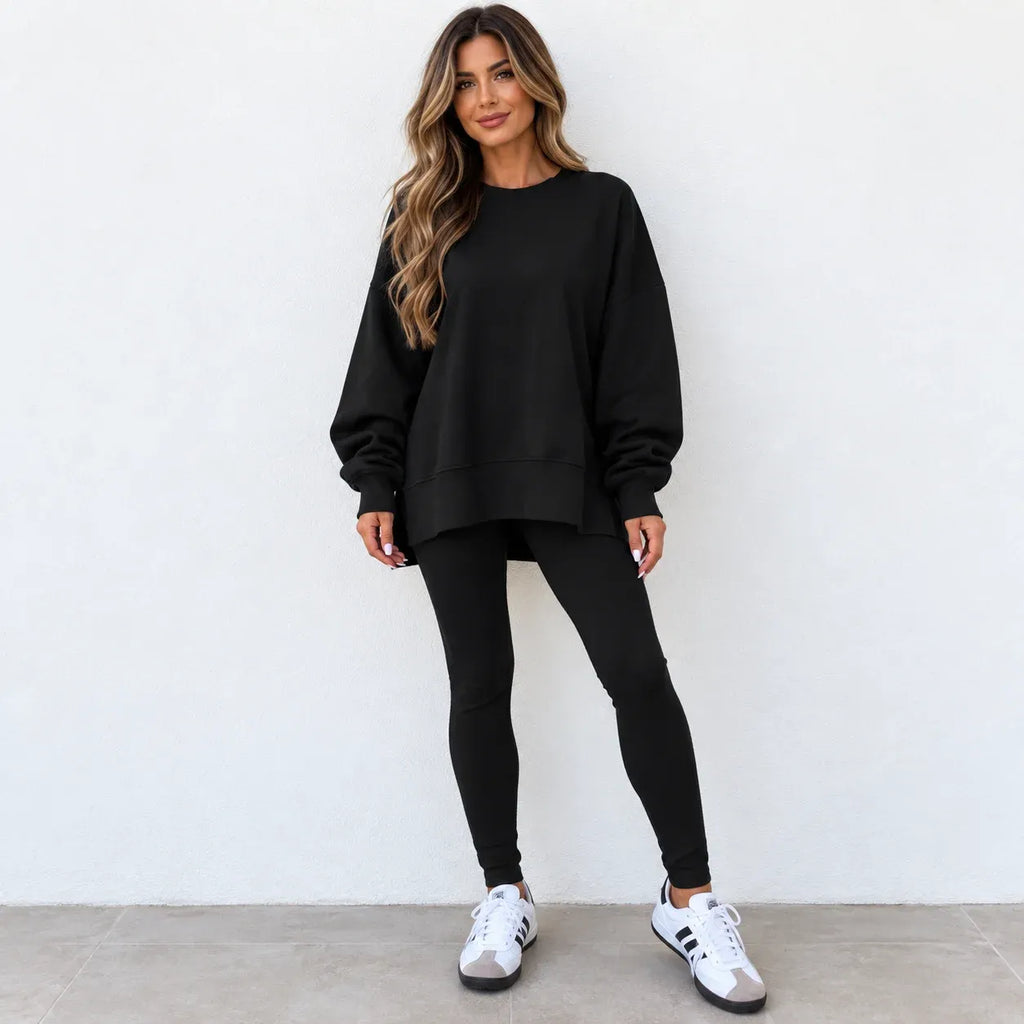 A woman modeling a black oversized crewneck sweatshirt with matching black leggings and white sneakers, standing against a plain white wall.