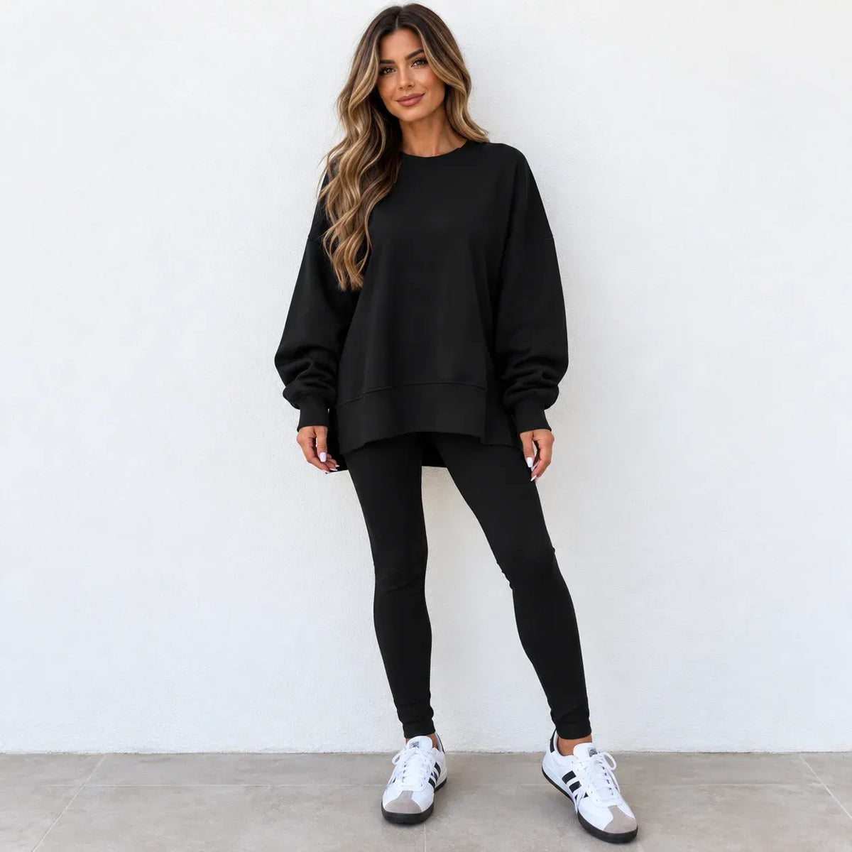 A woman modeling a black oversized crewneck sweatshirt with matching black leggings and white sneakers, standing against a plain white wall.