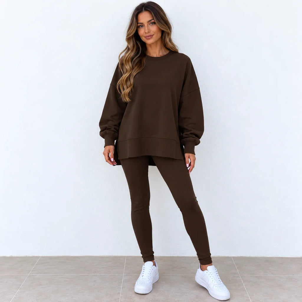 A woman modeling a matching dark brown lounge set consisting of an oversized long-sleeve sweatshirt and high-waisted leggings, paired with white sneakers against a plain white wall.