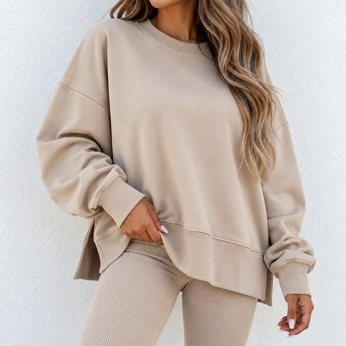 A woman wearing a beige crewneck sweatshirt and matching ribbed leggings, shown from the neck down against a light textured wall.