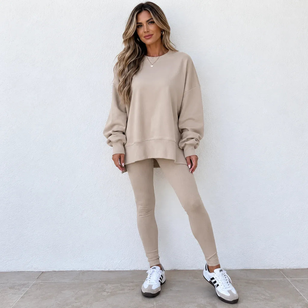 A woman modeling a matching beige lounge set consisting of a long-sleeve sweatshirt and high-waisted leggings, paired with white sneakers, standing against a white textured wall.